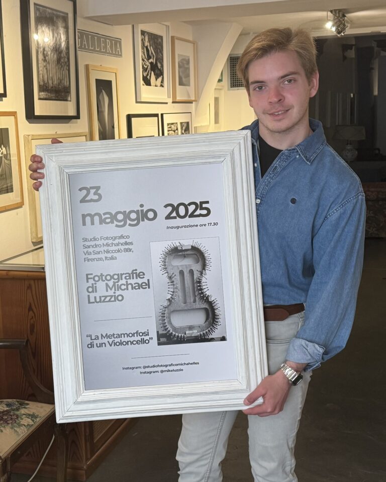 Michael Luzzio stands in an art gallery holding a large framed poster advertising his May 2025 photography exhibition in Florence.