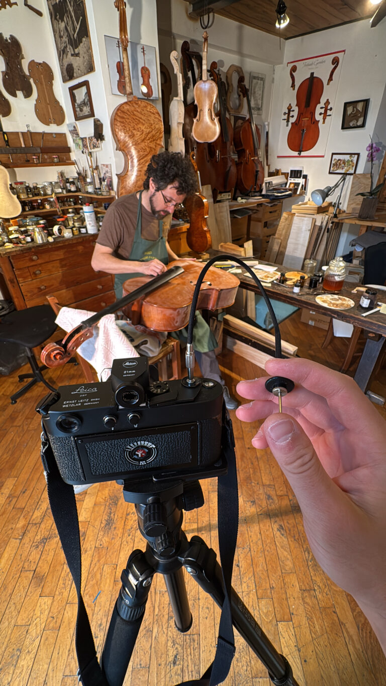 A hand holds a cable release on a tripod-mounted camera while a luthier works on a cello in a workshop filled with instruments and tools.