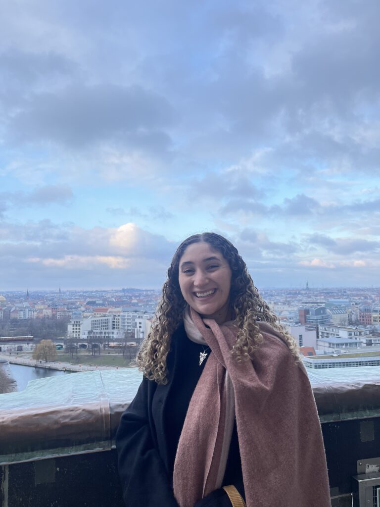 Paige Lovelace stands beside a red carpet in a bright, modern Brussels building, with geometric glass walls and EU flags visible in the background.