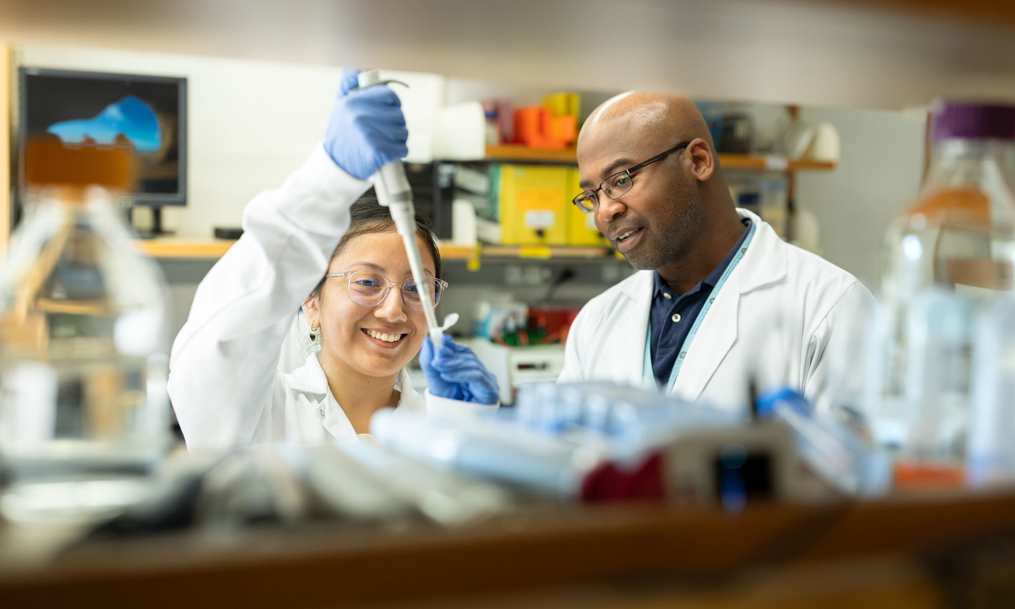 Two scientists in lab coats and gear smile while using equipment.