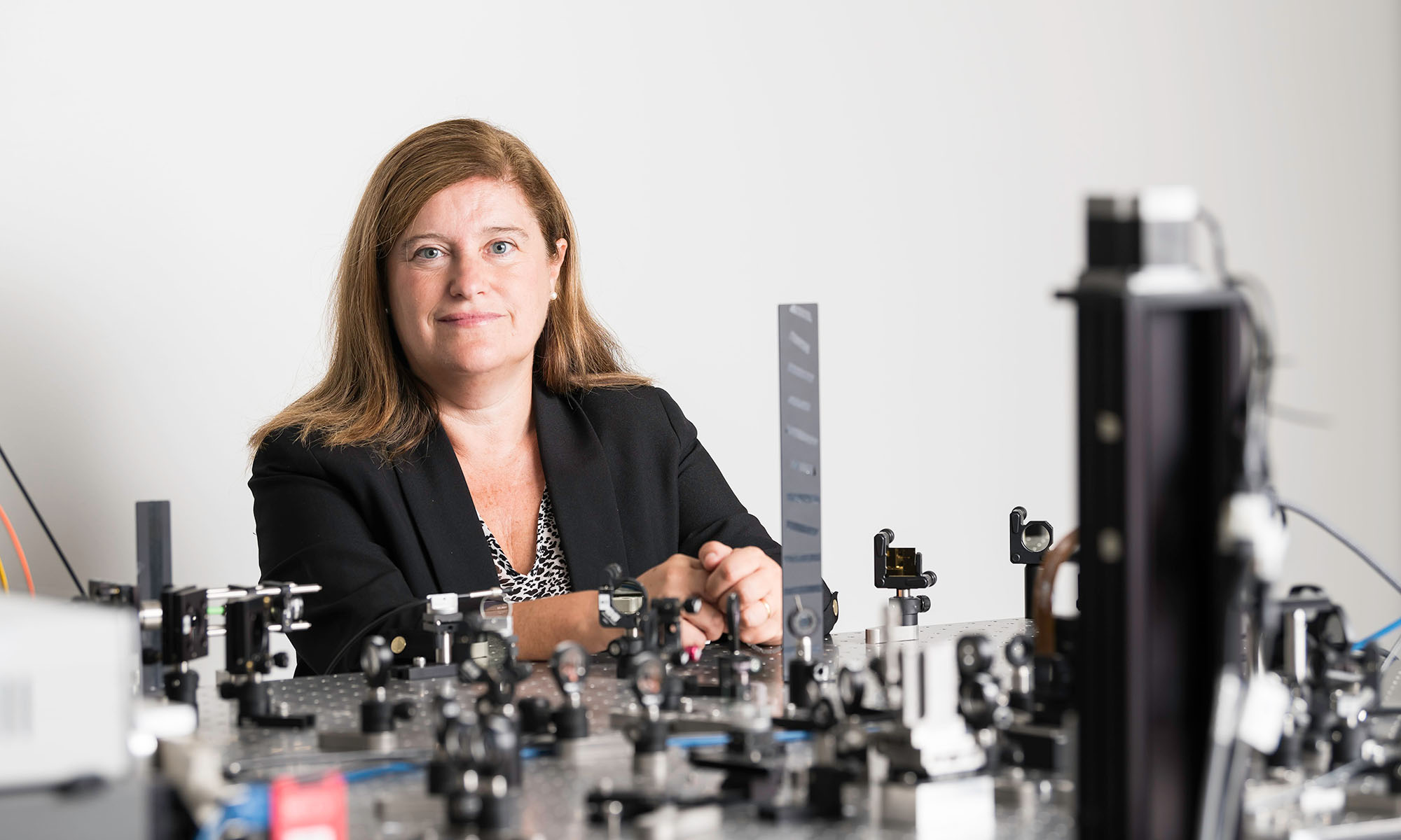 Susana Marcos smiles at the camera in her lab with the Center for Visual Science. In front of her is an optical array used for research.