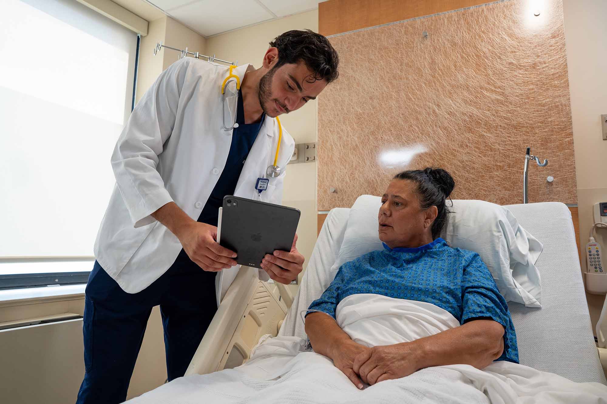 A physician wearing a white coat leans over a patient in a hospital bed to review their chart. 