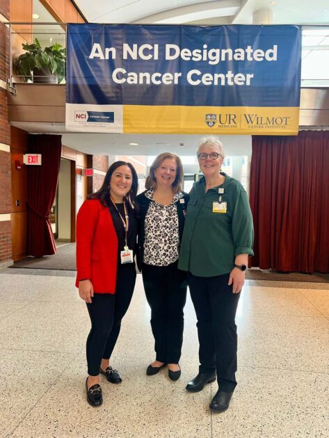 Lauren Ghazal, Lisa Kitko, and Sally Norton smile and stand together in front of a giant banner that reads "an NCI-designated cancer institute" and features the University of Rochester logo.