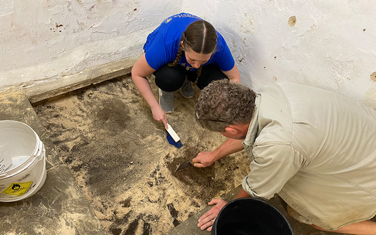 A student and instructor kneel on a dusty floor, carefully brushing and scraping during an indoor archaeological excavation activity.