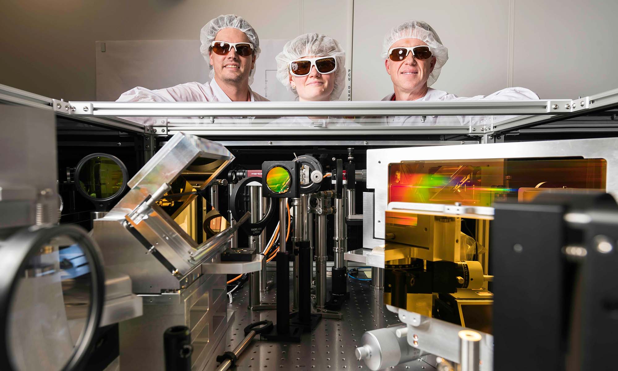 Three scientists in lab safety gear stand behind a laser array used to undertake chirped pulse amplification research at the Laboratory for Laser Energetics.