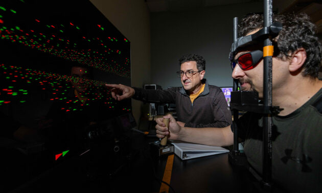 Two male researchers sit in a dimly lit office and look at a large computer screen displaying red and green chevrons in motion. One researcher is wearing an eye-tracking device.
