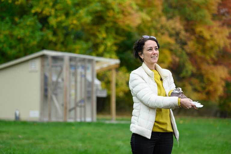 Dora Biro smiling and holding a homing pigeon with the pigeon coop in the background.