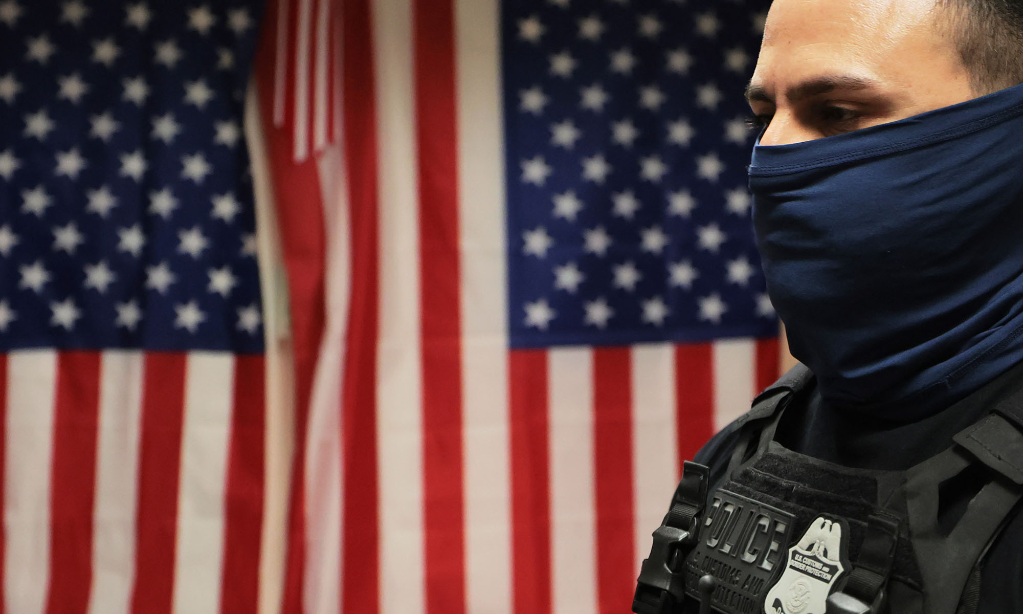 An Immigration and Customs Enforcement (ICE) agent with a mask covering his face stands in front of two American flags.