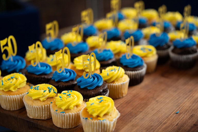 Rows of cupcakes with alternating blue and yellow frosting and 50th cake decorations on them.