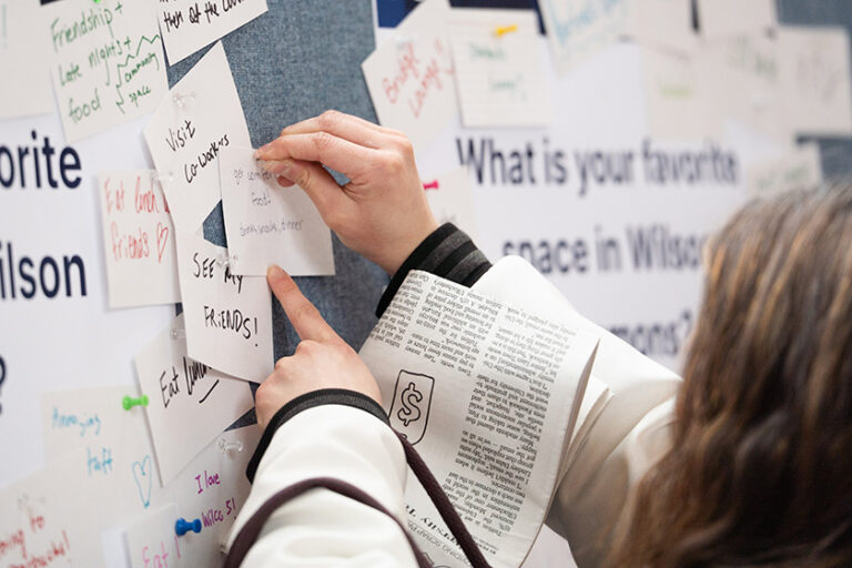 Close up of a students handings pinning a card to a mounted sheet asking about favorite spaces in Wilson Commons.