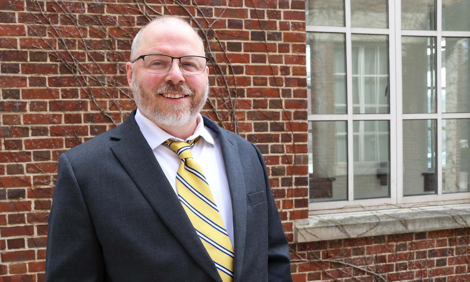 Greg Jorgensen in a suit smiling at the camera in front of a brick building on the URochester River Campus.