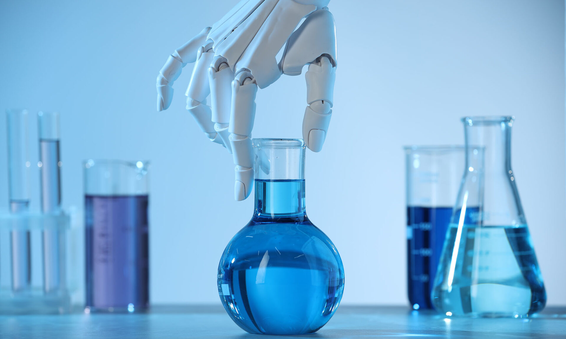Robot hand preparing to lift a flask of blue liquid with beakers and tubes of other liquids in the background.