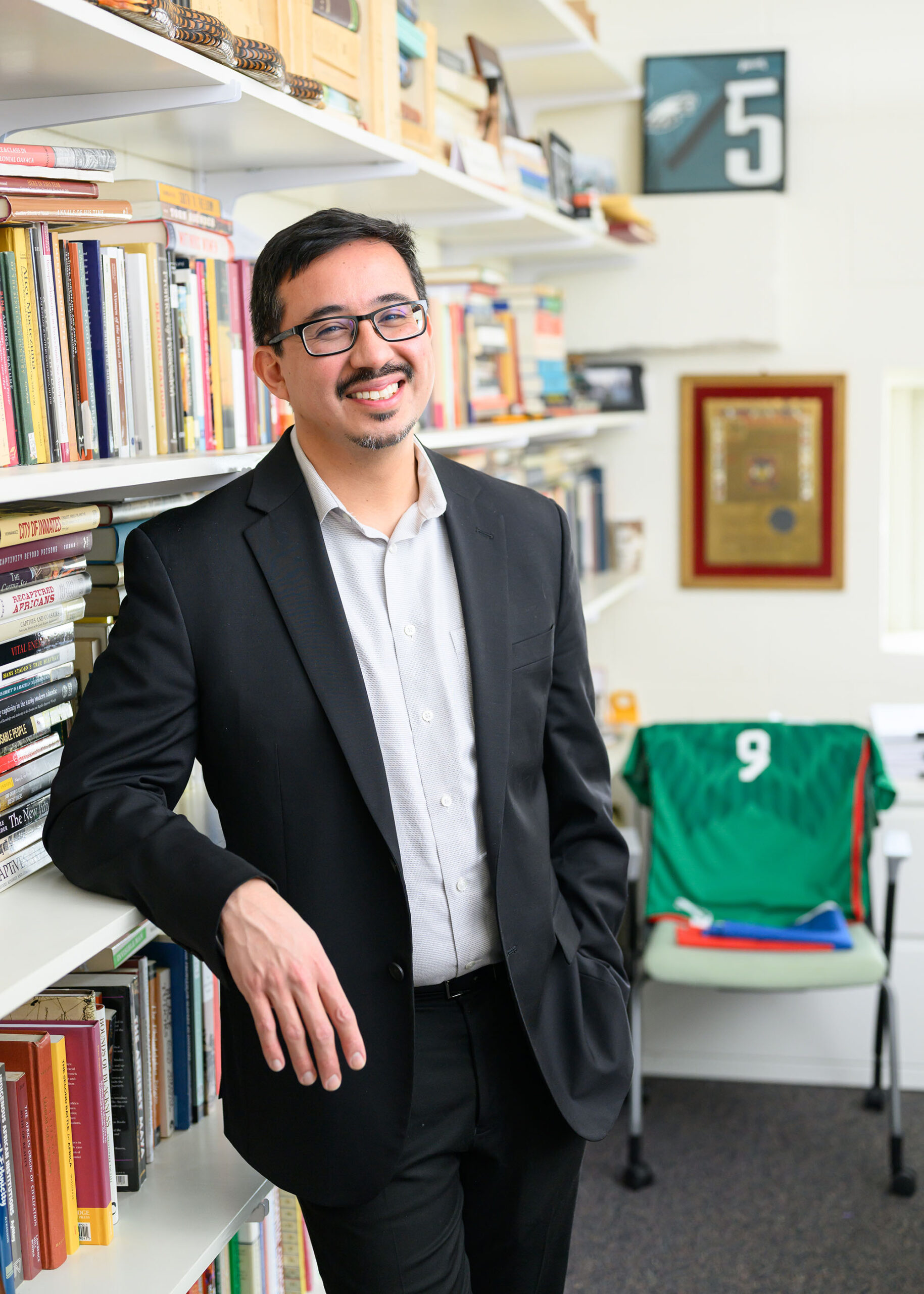 Pablo Sierra Silva leaning against a bookshelf in his office, smiling, with a soccer jersey and sports memorabilia visible behind him.