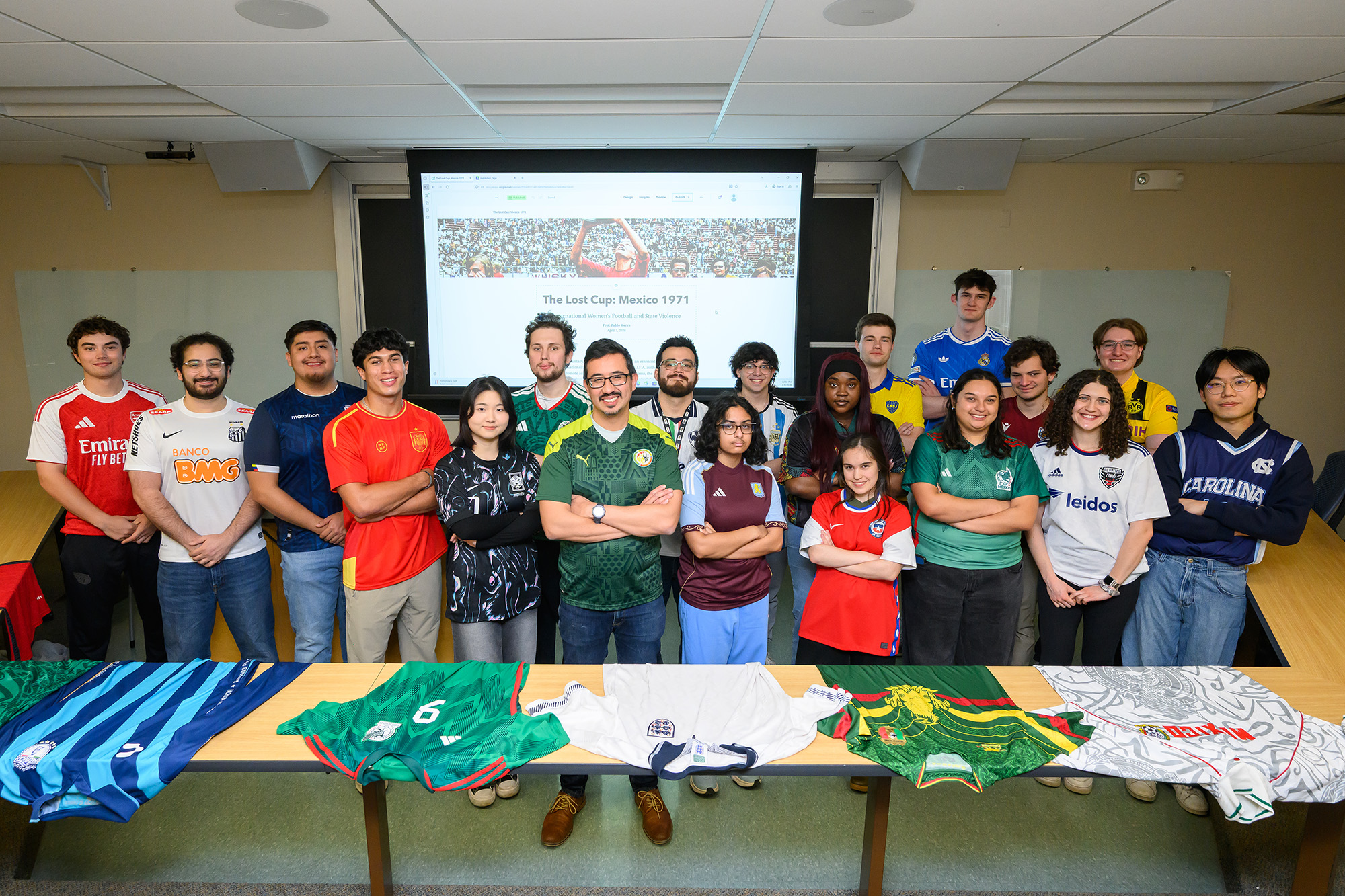Pablo Sierra Silva and his World History Through Soccer students pose in soccer jerseys in front of a projected lecture slide.