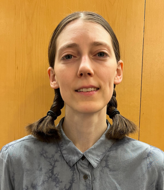 A woman with dark hair in two braids smiles at the camera in a professional headshot, wearing a gray patterned shirt against a wood-paneled background.