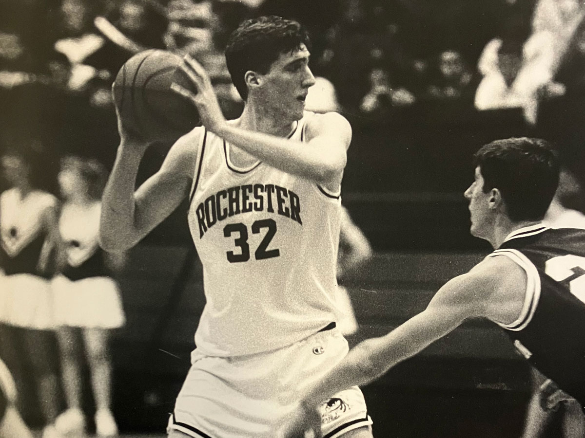 A black and white photo of a man in a white uniform holding a basketball, representing the University of Rochester.