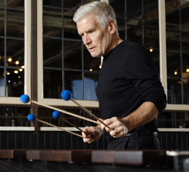 A man in a black long-sleeve shirt plays a marimba using four mallets with blue felt heads, captured mid-performance.