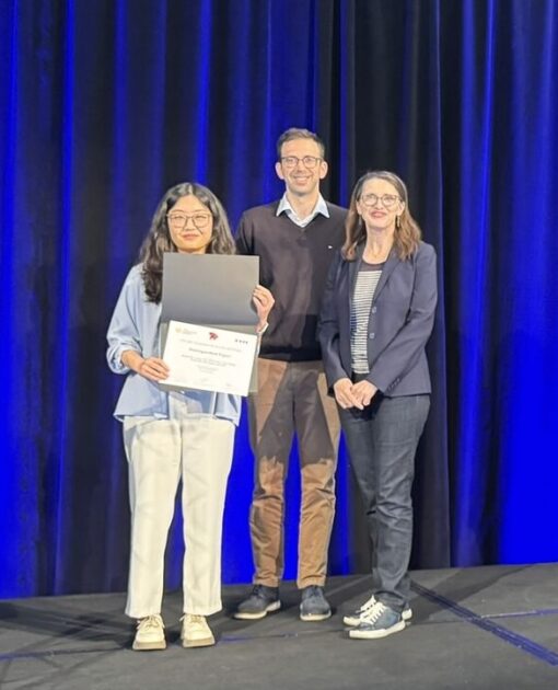 Three people pose together on a stage in front of a blue curtain, with one person holding an award certificate in a black portfolio folder.