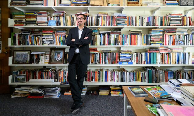 Pablo Sierra Silva stands with arms crossed in his Rush Rhees office in front of full bookshelves. (University of Rochester / J. Adam Fenster)