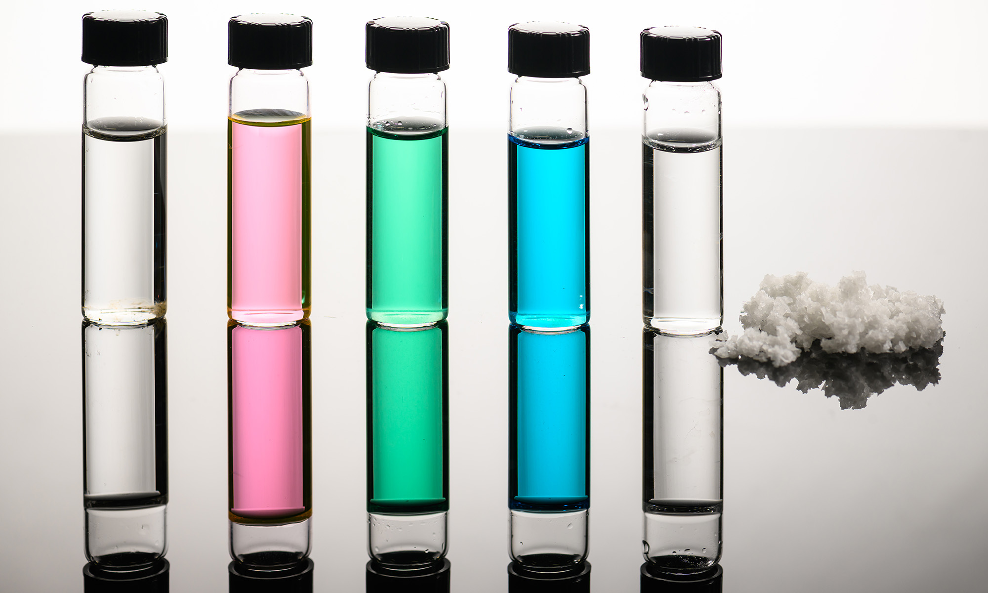 Five different colored vials holding seawater, Great Salt Lake water, nickel sulfate, copper chloride wastewater, and desalinated water standing next to a pile of salt.