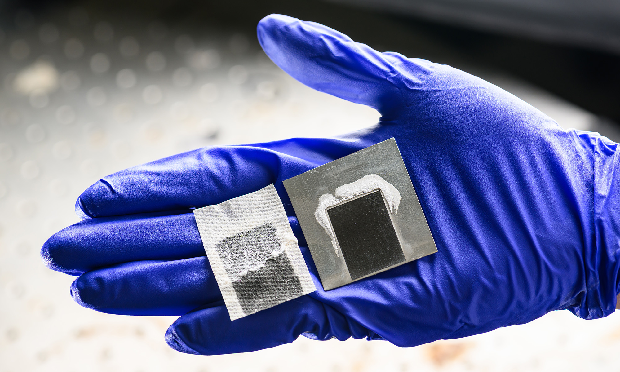 A blue gloved hand holding two pieces of laser-etched superwicking black metal.