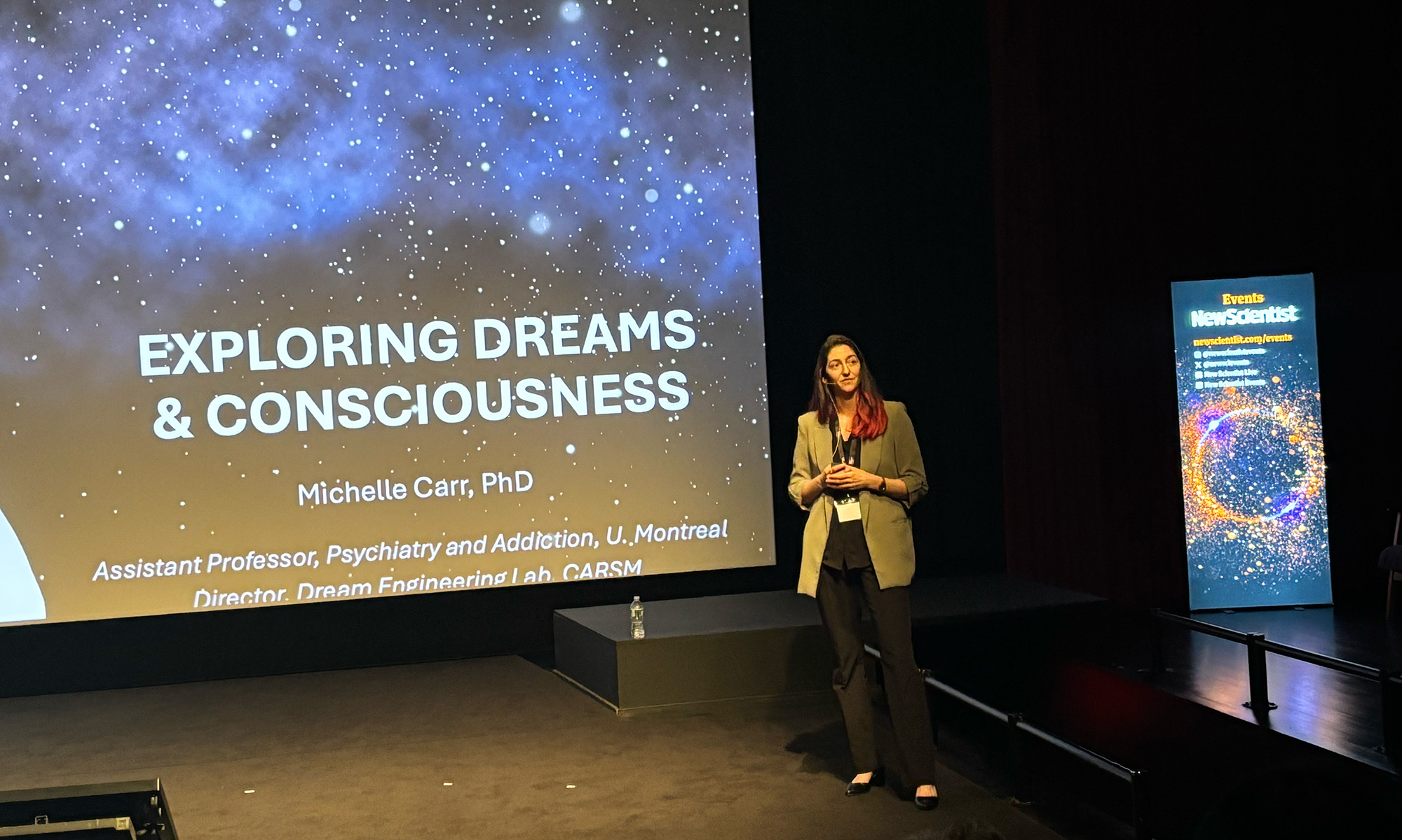 Michelle Carr presenting on stage at a New Scientist event, with a slide reading "Exploring Dreams & Consciousness" behind her.