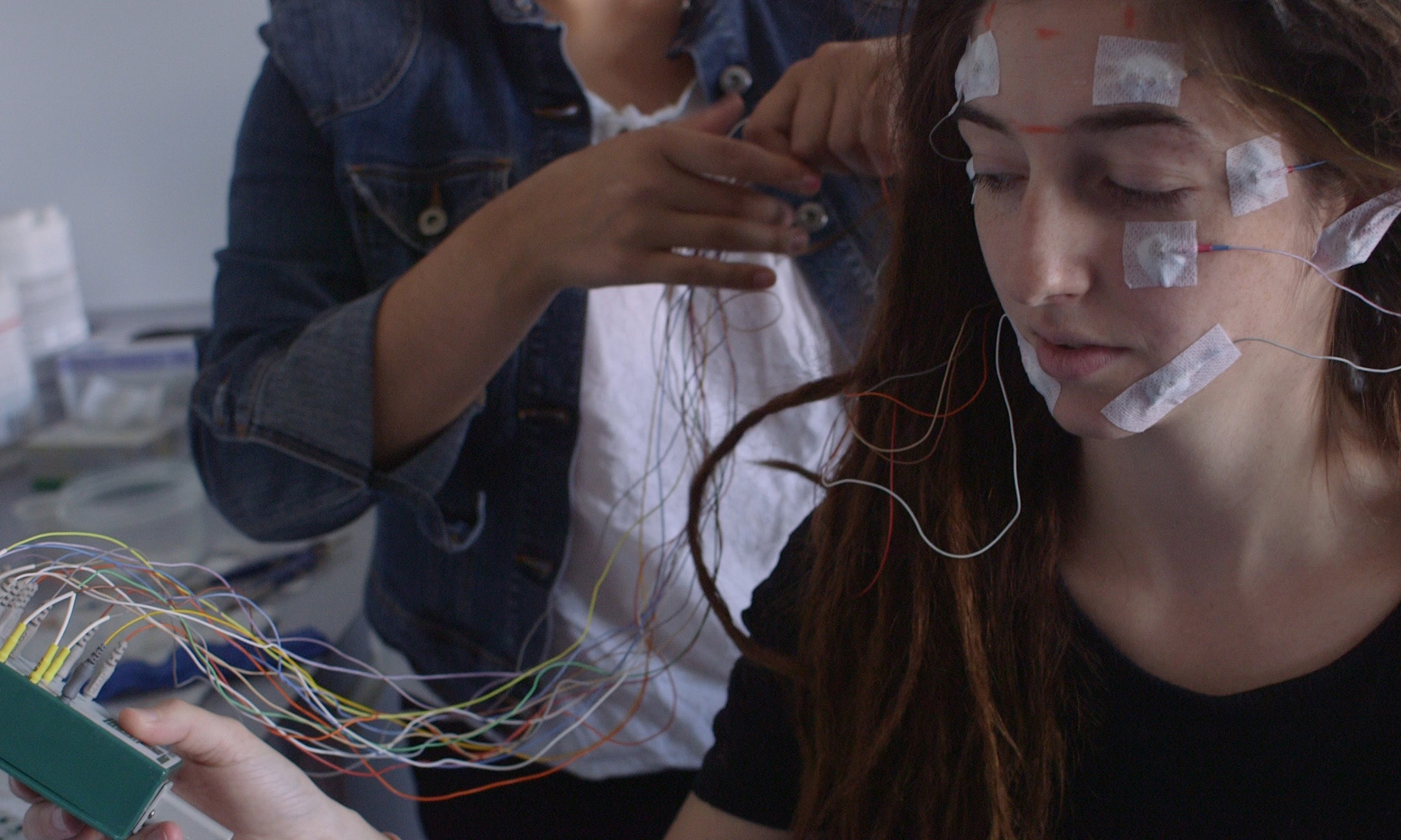 A researcher attaches colorful electrode wires to a study participant's face and head in a sleep research lab.