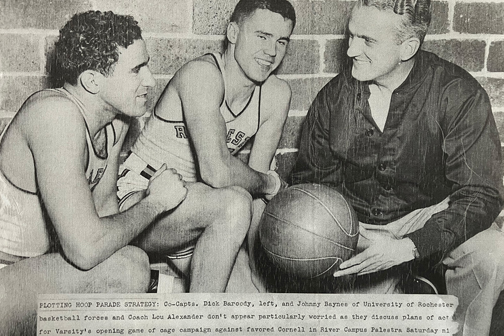 Co-captains Dick Baroody and Johnny Baynes confer with Coach Lou Alexander over a basketball in a black-and-white photo.