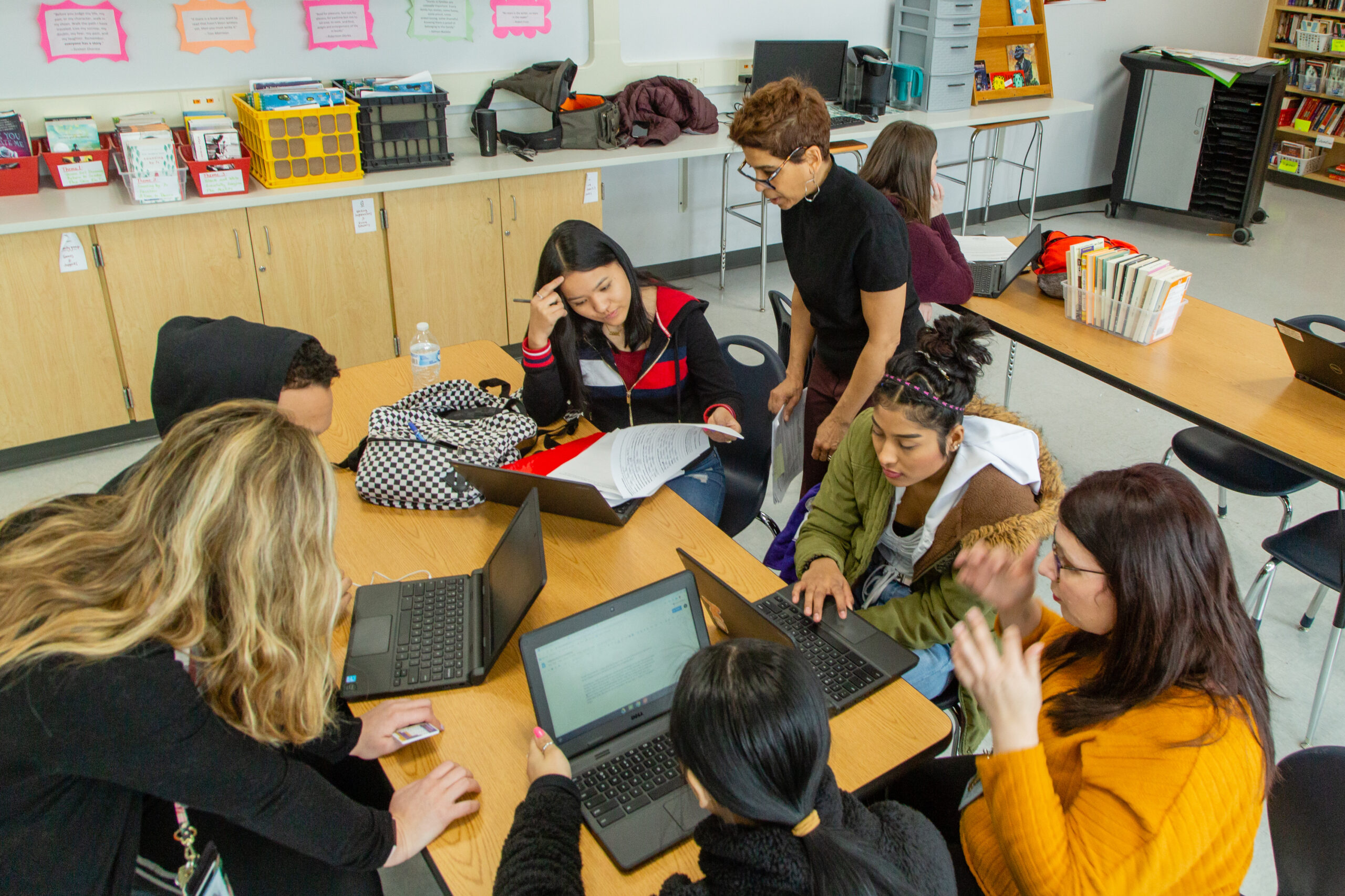 A group of high school students sits in a classroom at a table with laptops and papers while teachers help them with their work.