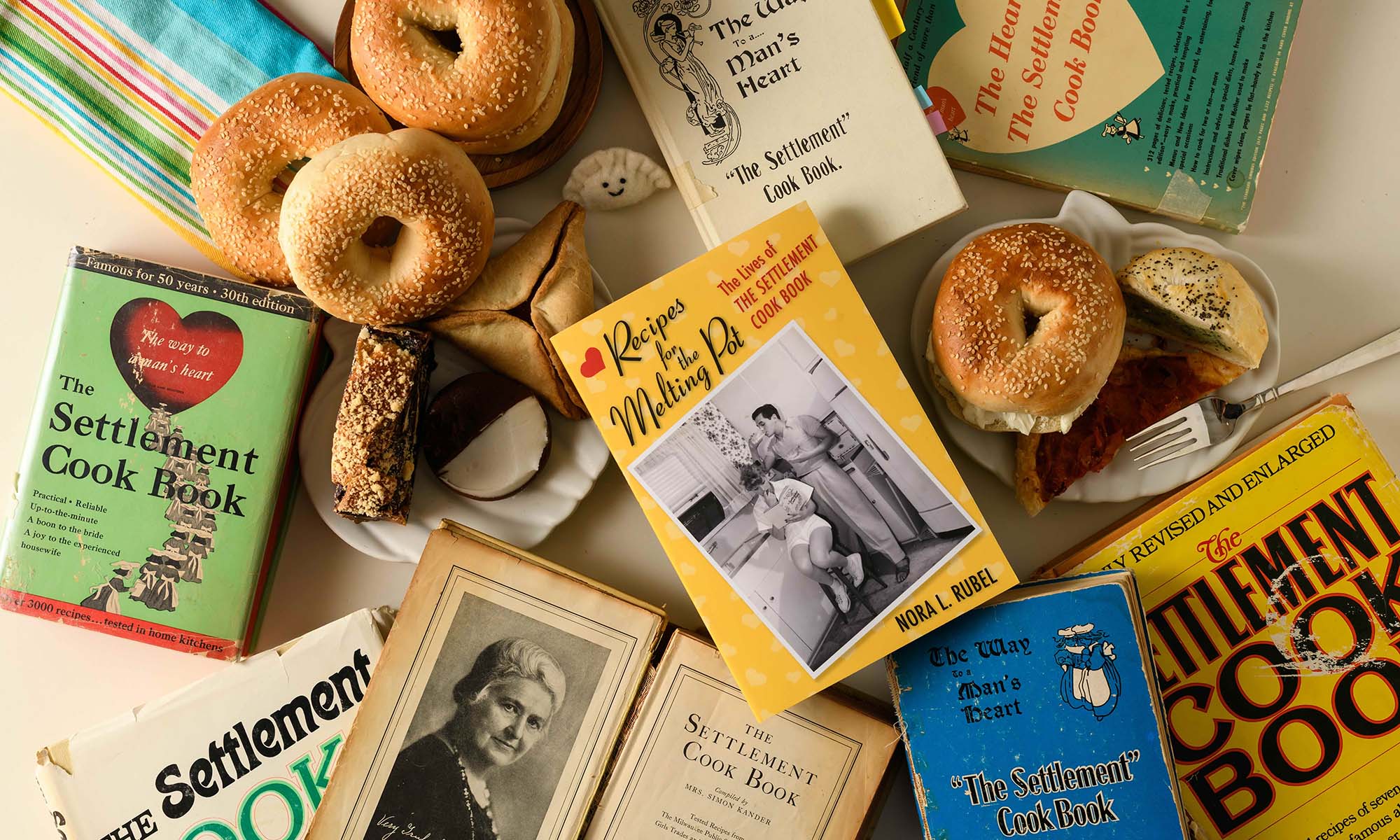 Overhead shot featuring several editions of The Settlement Cook Book alongside Jewish foods like bagels, babka, and hamantaschen, with the central focal point on Nora Rubel's book, "Recipes for the Melting Pot: The Lives of the Settlement Cook Book."