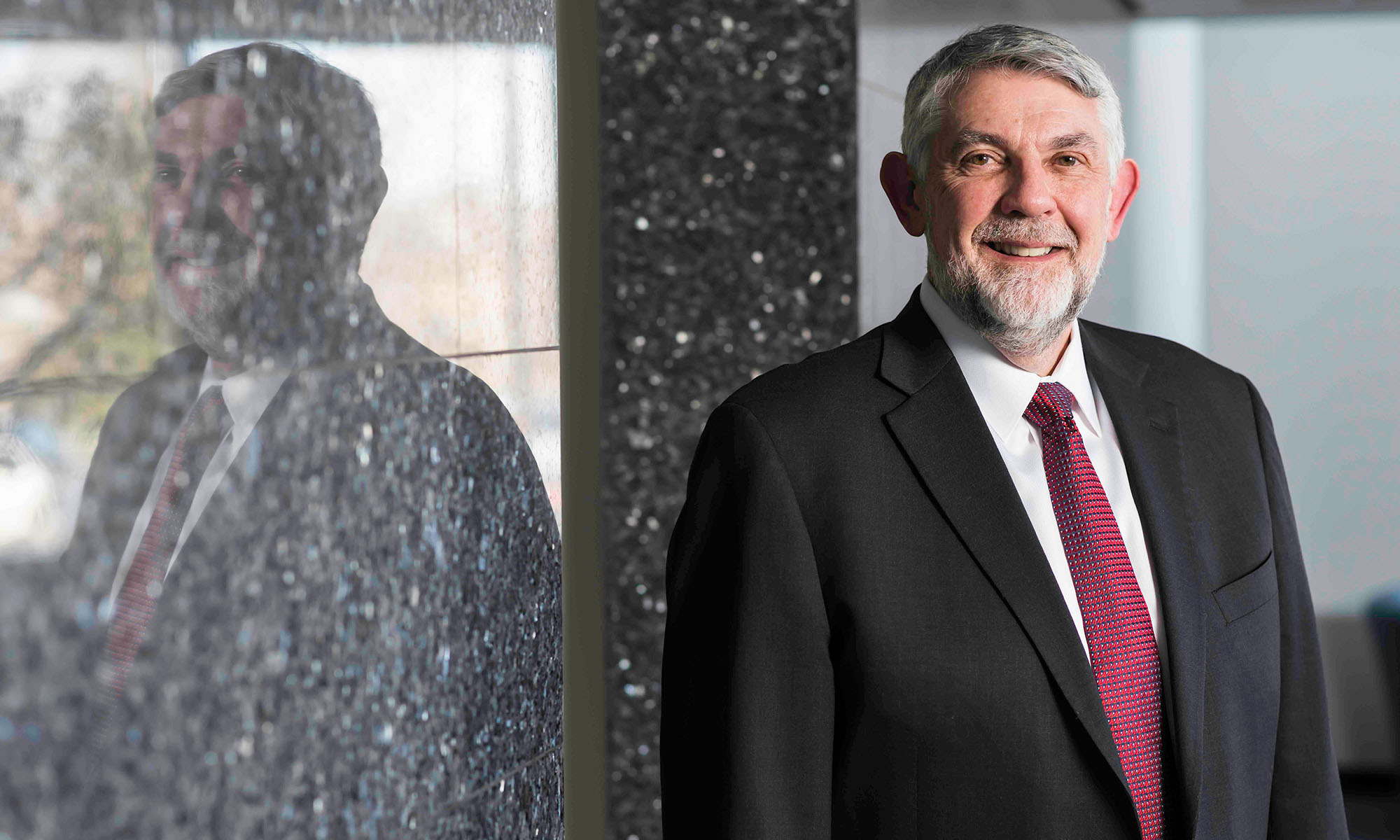 Christopher Deeney in a suit and tie with his image reflected on the wall next to him.
