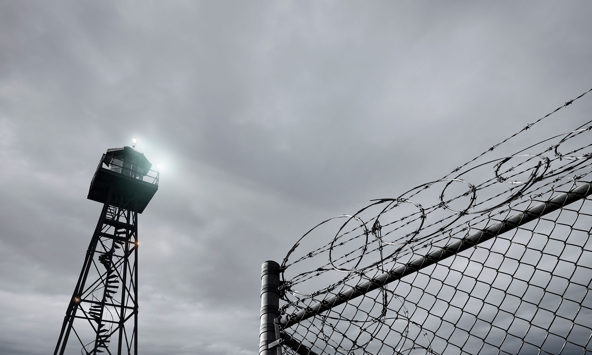 Black and white photo of a ecurity tower and razor wire-topped fence to represent incarceration in a prison or jail.