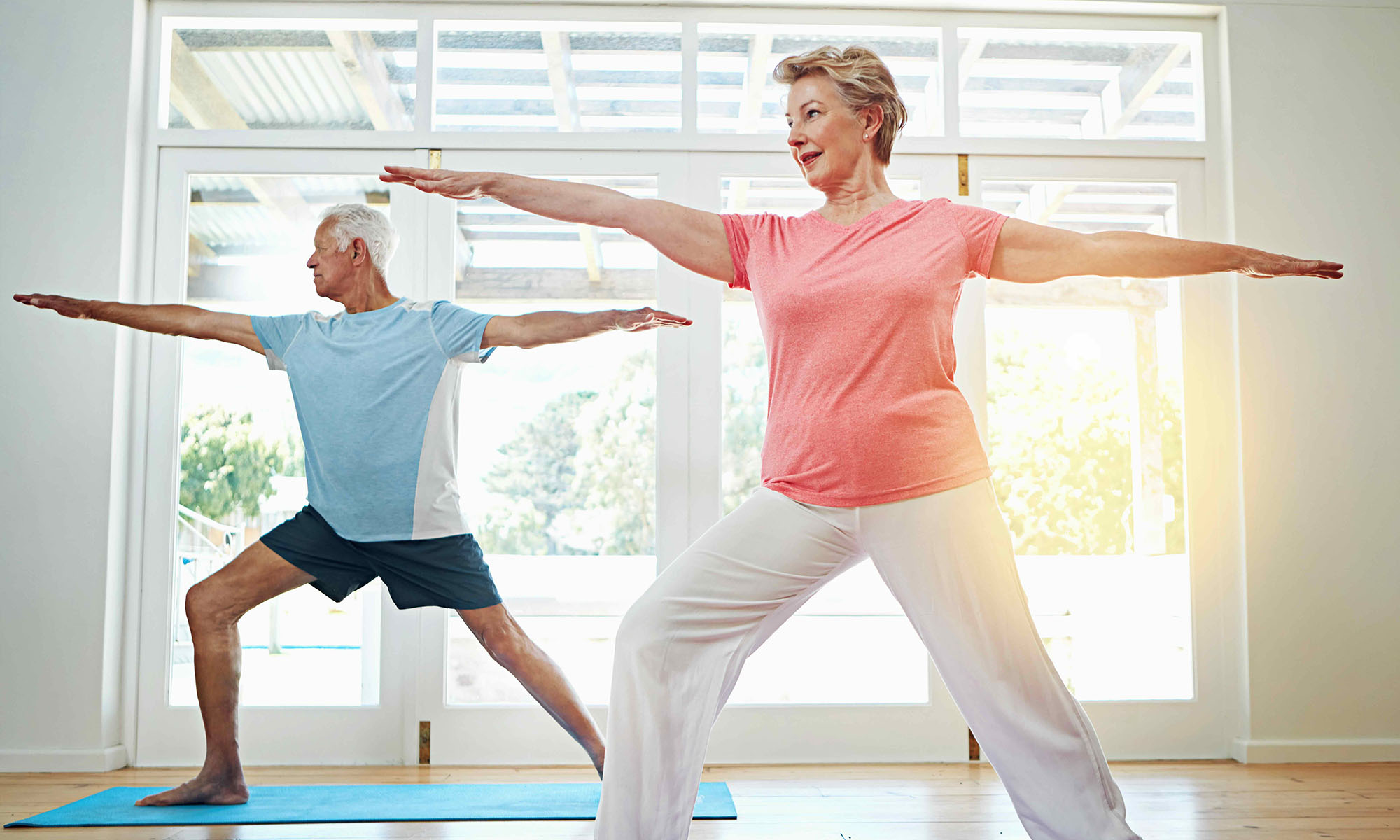 An elderly man and woman do yoga stretches in a sunlit room.