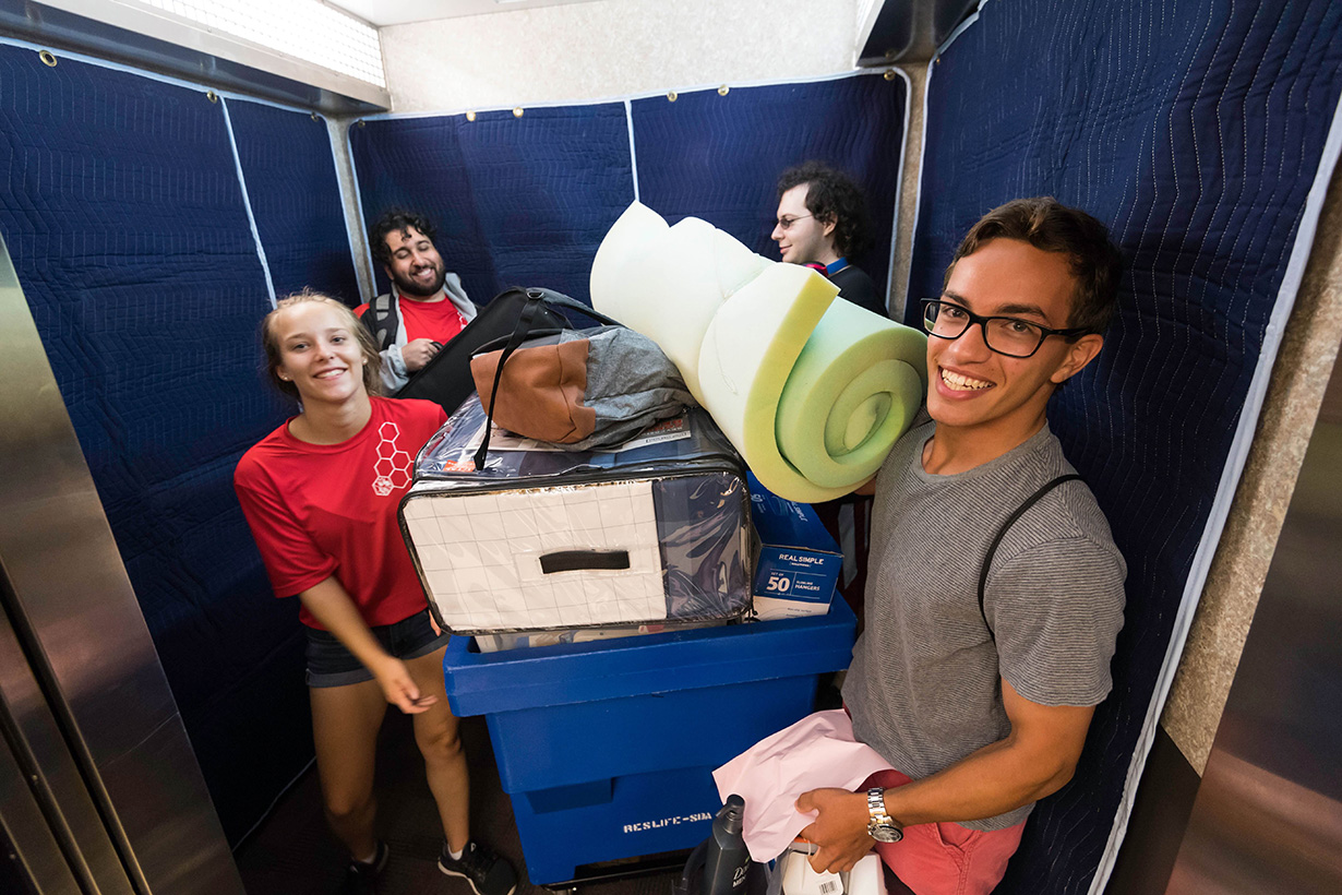 Four people in an elevator moving in the essential college packing list items for dorm life.