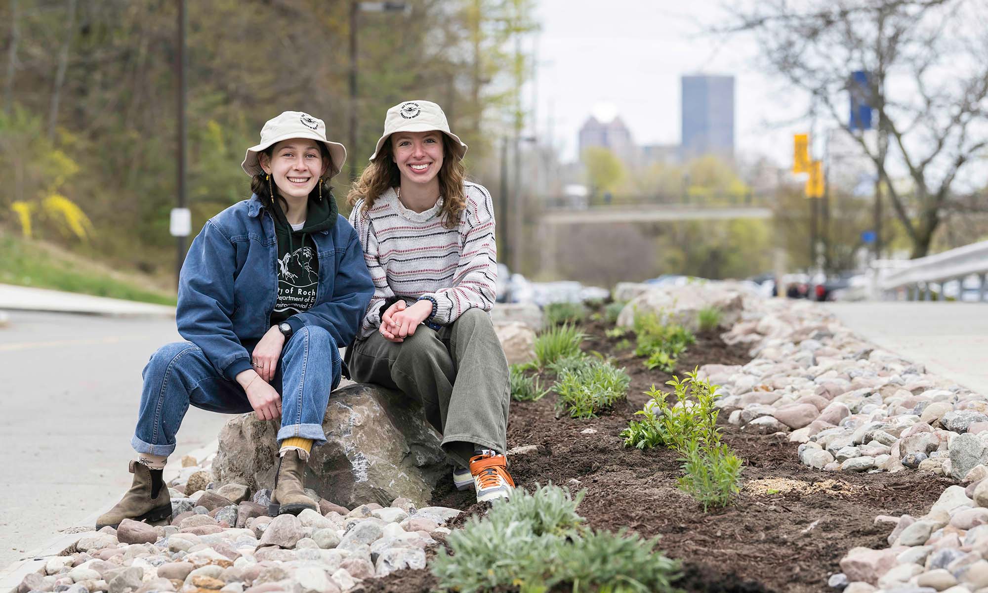 Two students in work clothes and sun hats sit beside a newly planted pollinator garden on the University of Rochester campus.