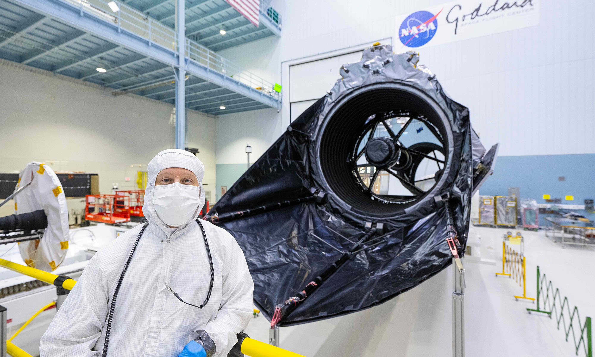Matthew Bolcar in a bunny suit stands beside NASA’s Nancy Grace Roman Space Telescope inside a clean room.