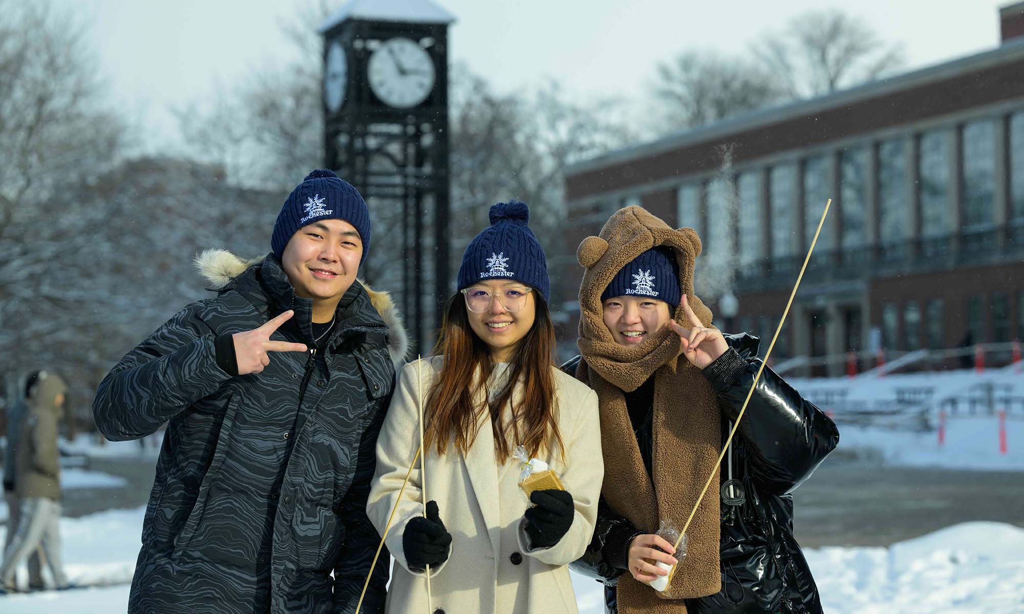 Three URochester students in winter garb smile for the camera in front of the Clock Tower.