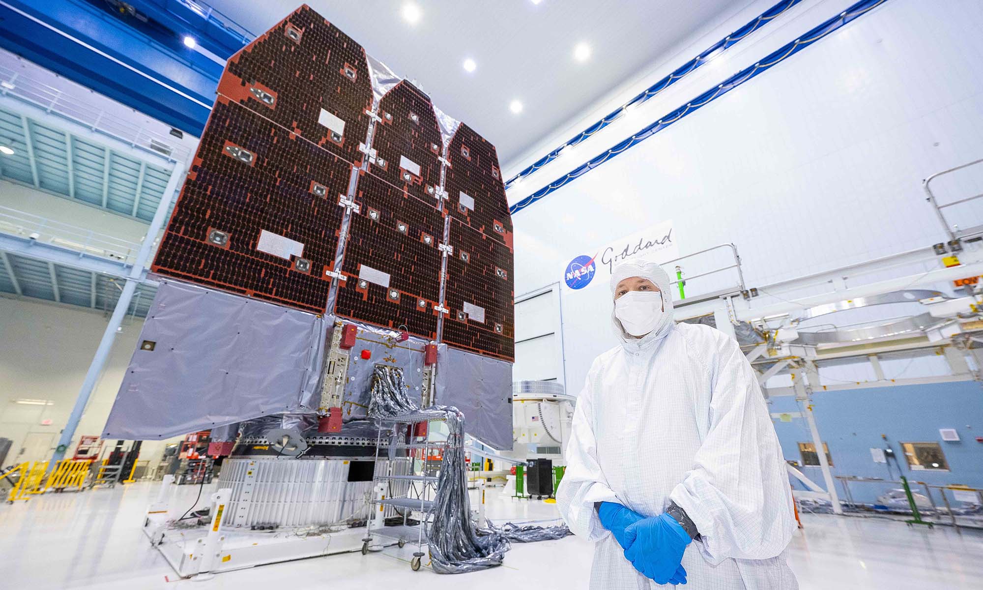 Alden Jurling stands in a bunny suit inside a clean room with NASA’s Nancy Grace Roman Space Telescope behind him.