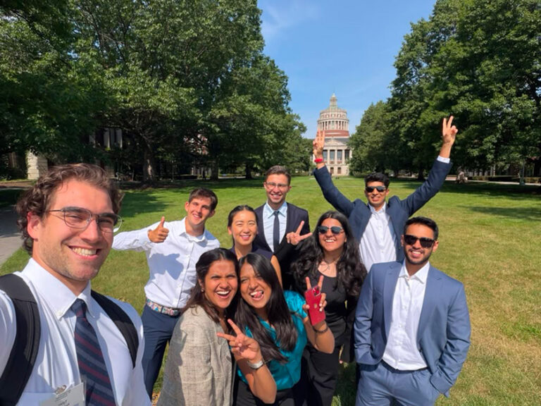 Group of Simon MBA students smile for a group selfie on the Eastman Quad with Rush Rhees Library in the background.
