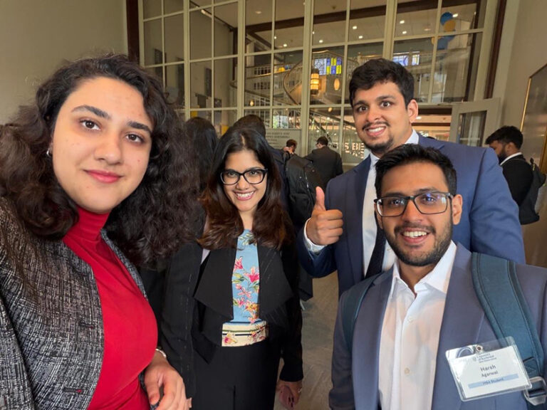 Four Simon Business School MBA students in business dress smile for a group selfie inside a campus building.