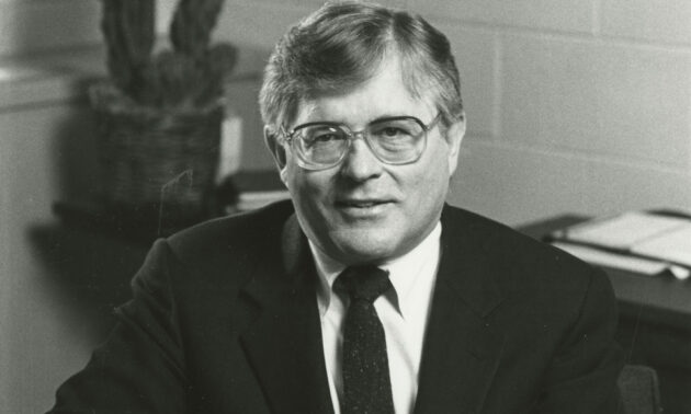 Horizontal crop of a black-and-white archival photo of Nicholas George in a suit in an office.