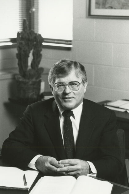Black-and-white archival photo of Nicholas George in a suit seated at desk with books open in front of him.