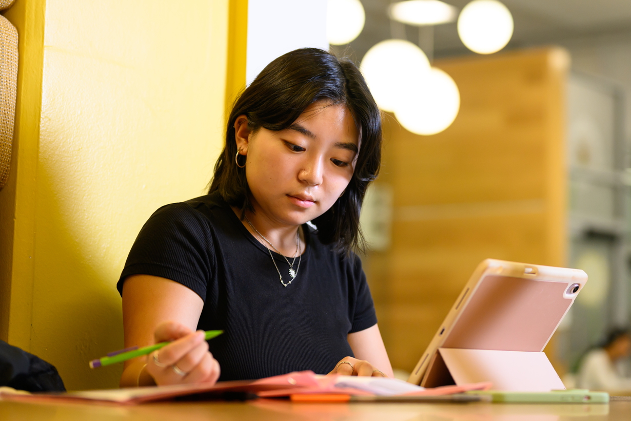 A URochester student looks down at her notebook while writing at a table that also has her phone nearby and tablet propped up.