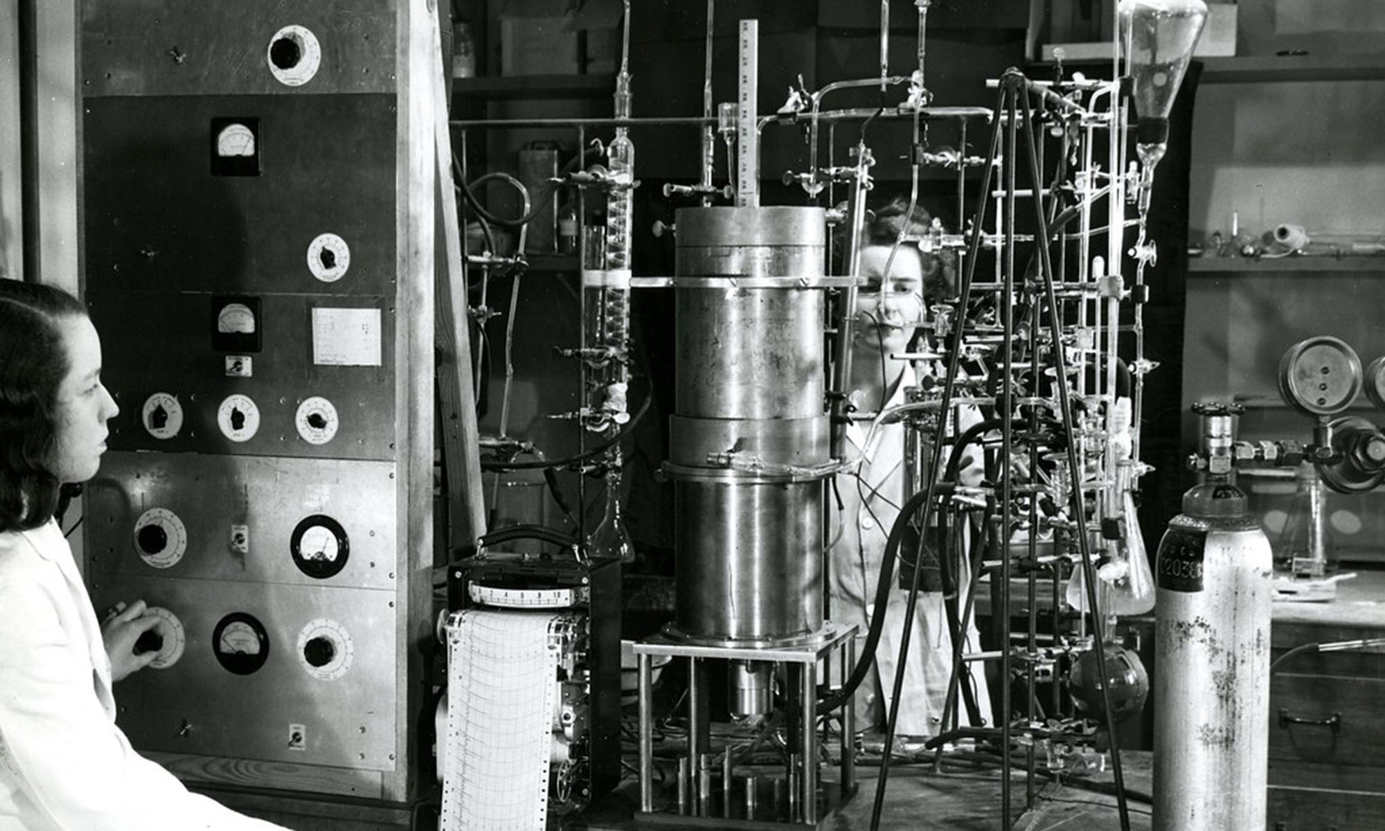 Black and white archival image of two researchers in white lab coats working with a complex array of laboratory equipment, glass tubing, gauges, and control panels.