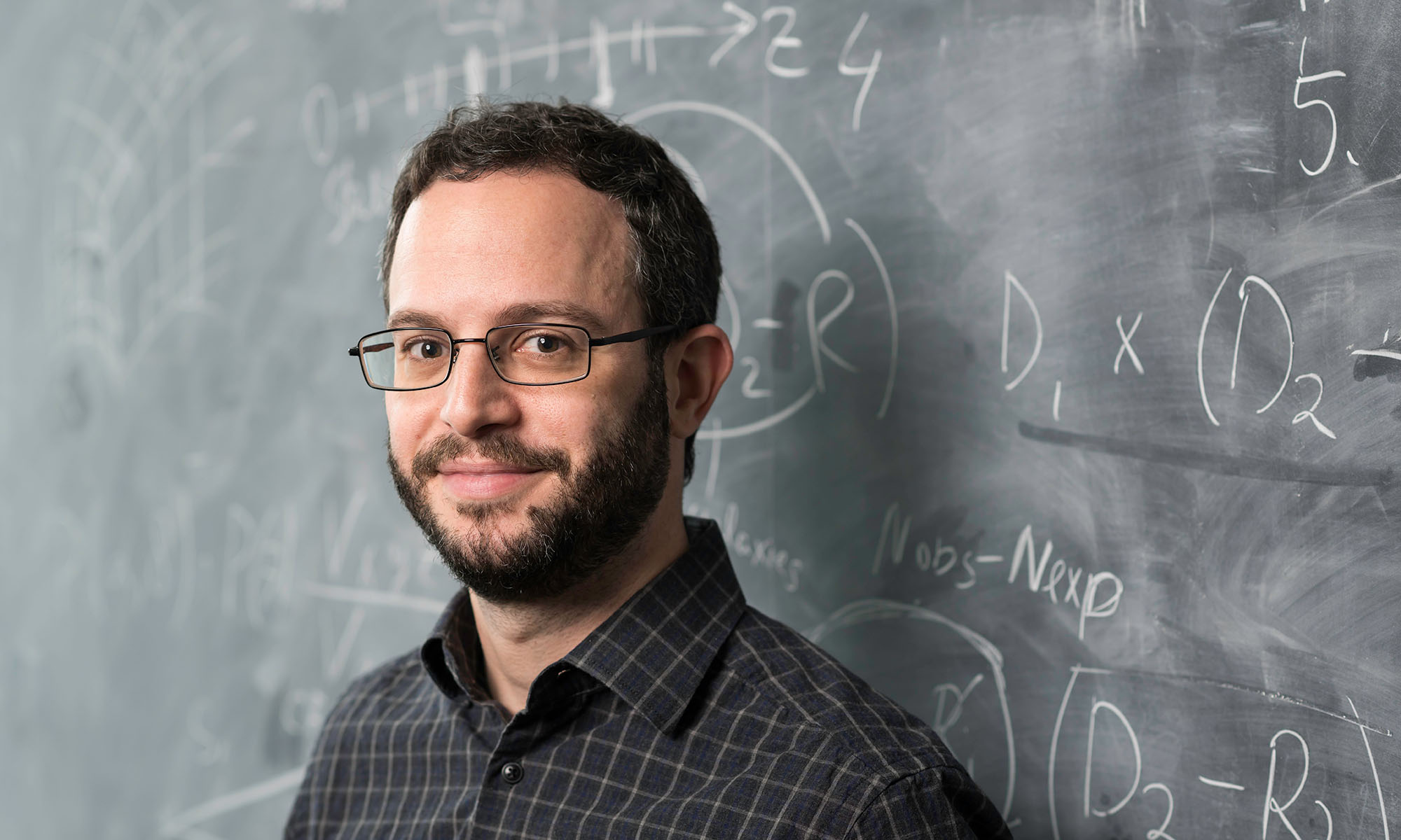 Segev BenZvi, who was a member of the team at the IceCube Neutrino Observatory, leans against a chalkboard with scientific equations on it.