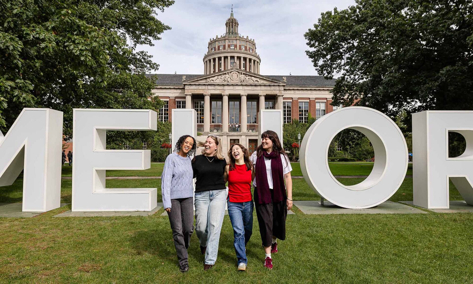 Four URochester alumni walking together in front of the MELIORA letters set up in front of Rush Rhees Library.