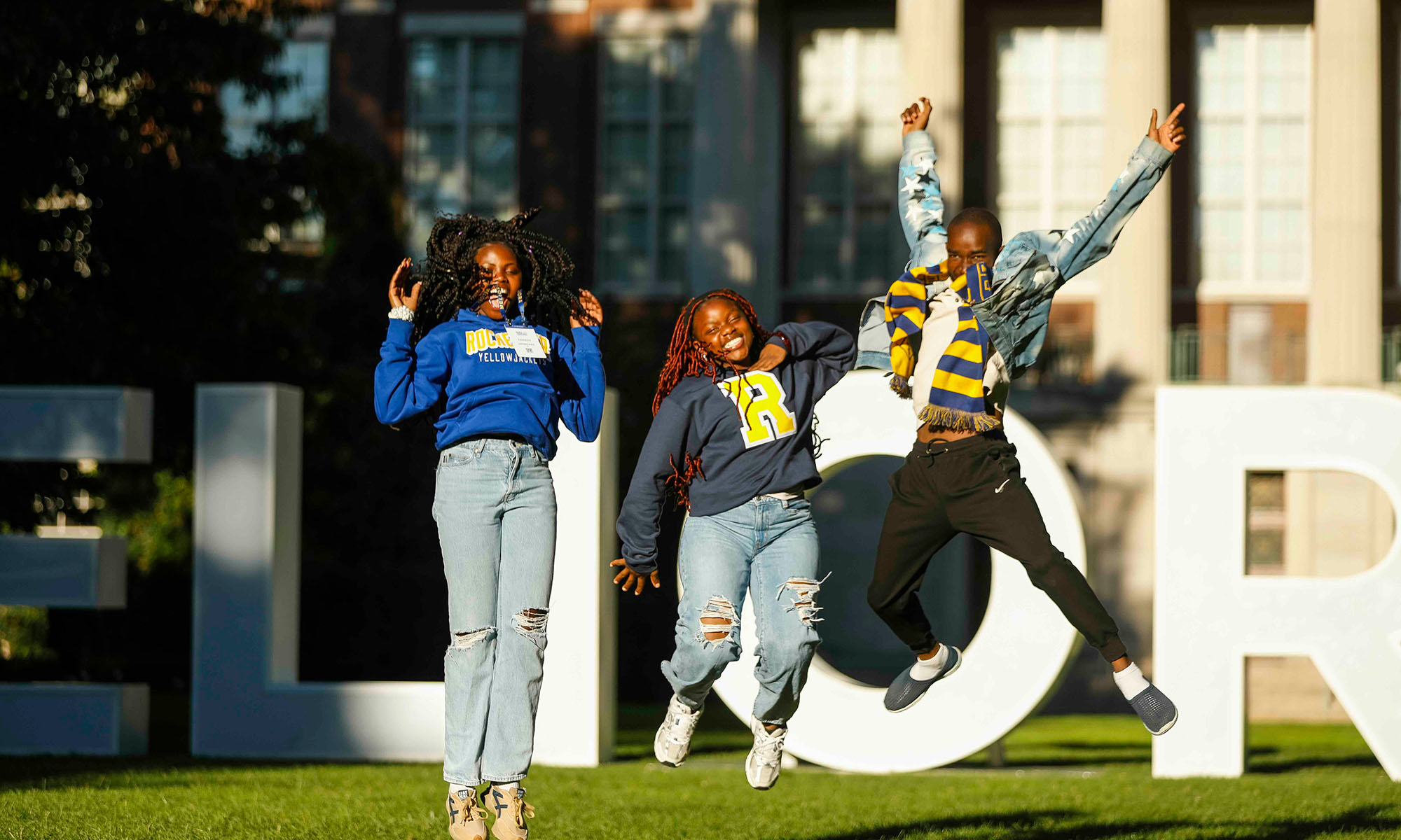 Three URochester students smiling and jumping in front of the MELIORA letters on the Eastman Quad.