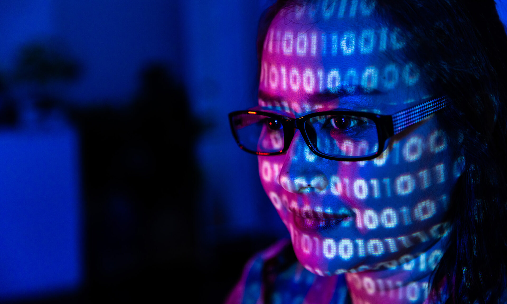 The face of a women wearing glasses bathed in blue and pink shadows and covered with binary code.