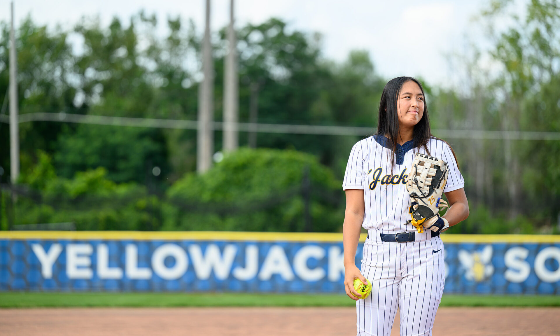 Riley Laygo stands in a Yellowjackets softball uniform holding a baseball glove and ball while standing on a baseball field.