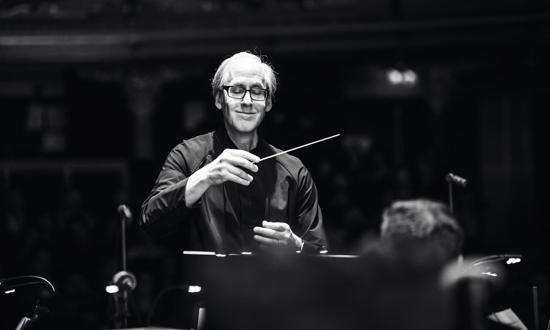 Composer Jeff Beal smiles while conducting, holding a baton in a black-and-white portrait.